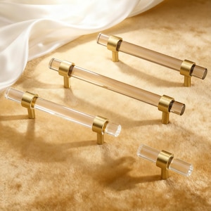 May include: Four cabinet pulls with clear acrylic rods and gold-toned metal accents. The pulls are arranged on a textured, golden-colored surface. The acrylic rods have a slight amber tint, and the metal hardware has a polished finish.
