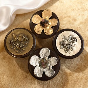 Może przedstawiać: Cztery dekoracyjne gałki do szafek z ciemnobrązowymi podstawami. Dwie gałki mają wzory motyli i róż, a pozostałe dwie wzory kwiatowe. Metalowe akcenty są w odcieniach złota i srebra.