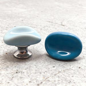 Puede incluir: Dos tiradores decorativos para armarios. Uno tiene forma de hongo, de color azul claro, montado sobre una base plateada. El otro es un tirador liso, de forma ovalada, en color verde azulado intenso. Ambos tienen un acabado brillante.