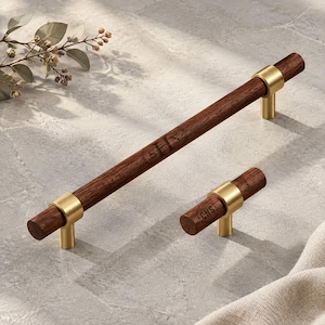 May include: A long cylindrical dark wood cabinet pull and a smaller matching T-shaped knob, both featuring brushed gold metal accents. The wood surfaces are engraved with the letters CNS inside a circular laurel wreath design detail.