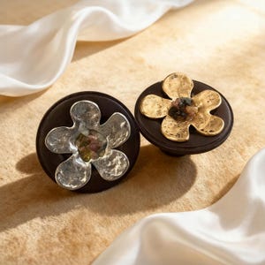 May include: Two decorative cabinet knobs. One knob features a silver flower design, and the other has a gold flower design. Both knobs are mounted on a dark brown circular base. The knobs are set against a neutral background with white fabric.