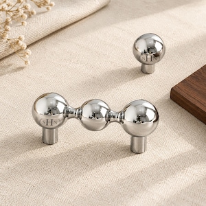 May include: A polished silver-toned metal cabinet pull featuring three connected spheres and a single matching round knob. Both pieces are engraved with the letters SHN inside a circular border. The hardware has a reflective, mirror-like finish.