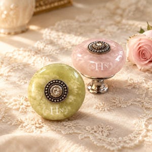 May include: Two round cabinet knobs, one in pale green and one in soft pink, featuring a marbled texture. Each knob has a central silver-toned metal accent with a small embedded crystal and the engraved letters SHN surrounded by laurel leaves.