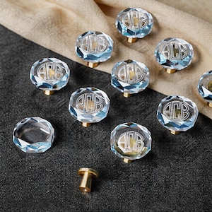 May include: A collection of faceted, light blue glass cabinet knobs with a circular gold-toned base. Each knob features a central engraved monogram with the letters ACB inside a circular border. One knob is shown detached, revealing the gold hardware.