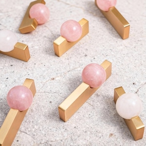 Puede incluir: Tiradores de armario de oro y cuarzo rosa. Cada tirador tiene una base rectangular dorada con una esfera de cuarzo rosa. Diseñados para armarios, cajones o muebles. Las esferas de cuarzo rosa varían de rosa claro a rosa medio.