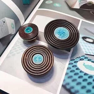 May include: Three decorative cabinet knobs with a concentric ring design in a dark brown finish. Each knob features a central, circular inlay of turquoise-colored ceramic. The knobs vary in size, suggesting they are for different cabinet applications.