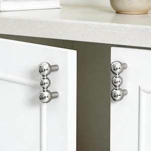 Peut inclure: Gros plan sur des portes d'armoires blanches avec des poignées de meubles argentées à trois sphères. Les poignées sont alignées verticalement et ont une finition polie et réfléchissante. Les portes sont épurées et modernes.