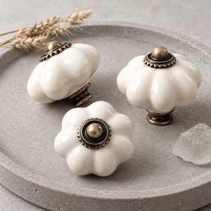 Puede incluir: Tres pomos de cerámica blanca para armarios con un diseño acanalado en forma de calabaza. Cada pomo cuenta con una base central de metal en tono latón antiguo y una tapa superior circular a juego con un borde dentado decorativo.