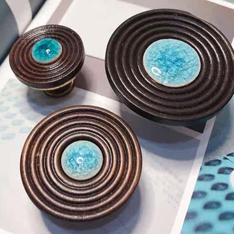 May include: Three decorative cabinet knobs with a concentric ring design. The knobs are made of dark brown wood and feature a central turquoise-colored, speckled glass or ceramic insert. One knob has a brass base.
