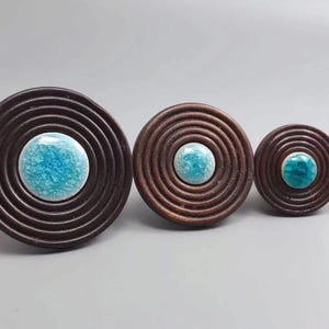 May include: Three decorative wooden knobs with concentric circle designs. Each knob features a central, round, turquoise-colored ceramic accent. The knobs vary in size, with the largest measuring approximately 3 inches in diameter.