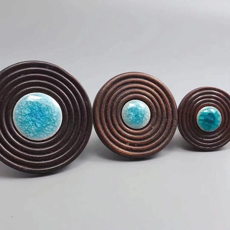 May include: Three decorative wooden knobs with concentric circle designs. Each knob features a central, round, turquoise-colored ceramic accent. The knobs vary in size, with the largest measuring approximately 3 inches in diameter.
