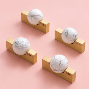 May include: Four cabinet knobs, each featuring a spherical white marble stone with grey veining set into a rectangular brushed gold metal base. Each base is engraved with the letters sHN in a small circular emblem on the right side.