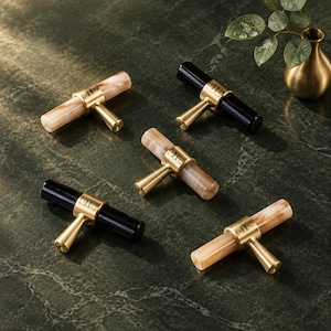 May include: Five T-bar cabinet knobs featuring cylindrical handles in black or marbled beige resin, each secured by a central gold-toned metal mount engraved with the letters SHN. The hardware pieces are arranged in a cluster on a dark surface.