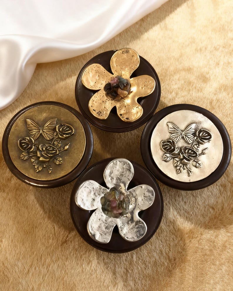May include: Four decorative round objects with dark brown bases. Two feature flower designs in gold and silver, while the other two have butterfly and rose motifs in gold and silver. The objects are arranged on a textured, tan surface.
