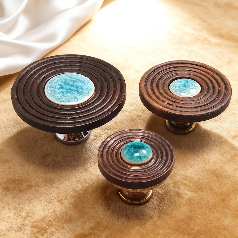 May include: Three decorative cabinet knobs. The knobs have a dark brown, circular design with concentric rings. Each knob features a central, round, light blue ceramic accent. The knobs have silver and gold hardware.