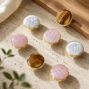 May include: A collection of seven circular cabinet knobs with gold-toned metal rims. Each knob features a unique inlay, including white marble, pink stone, and brown tiger's eye patterns. Every knob is engraved with the monogram letters ACB in the center.