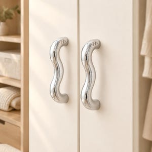 May include: Two polished chrome cabinet handles with a distinct wavy, serpentine shape. Each handle features a small, engraved SHN logo at the top. The handles are mounted vertically on a pair of smooth, matte off-white cabinet doors.