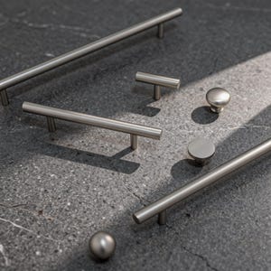 May include: A collection of brushed nickel cabinet hardware, including bar pulls, T-knobs, and round knobs. The hardware is arranged on a dark gray speckled surface, with shadows adding depth. The metal has a matte finish, reflecting soft light.