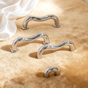May include: Several polished, silver-coloured cabinet pulls with a wavy design. The curved handles are displayed against a neutral background. These are likely for furniture or cabinetry, and are made of metal.