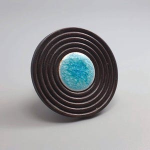 May include: A dark brown, circular cabinet knob with concentric rings. The center features a round, turquoise-colored ceramic insert with a crackle glaze. The knob is likely made of wood and ceramic.