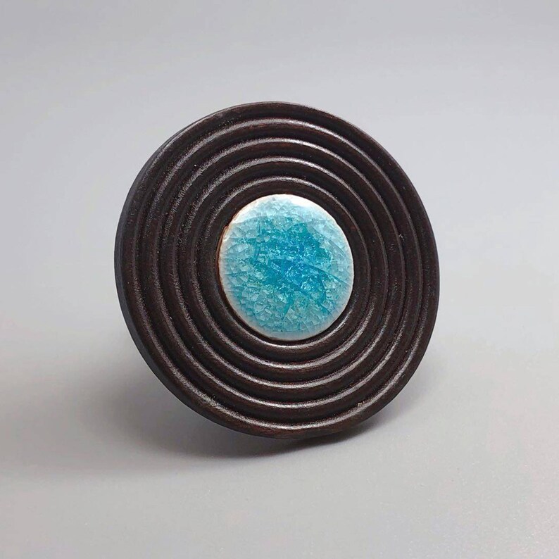 May include: A dark brown, circular cabinet knob with concentric rings. The center features a round, turquoise-colored ceramic insert with a crackle glaze. The knob is likely made of wood and ceramic.