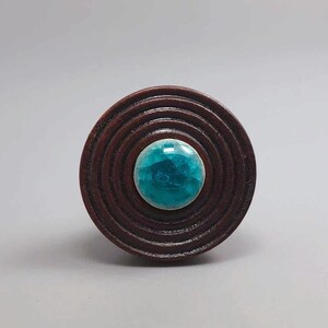 May include: A round, dark brown wooden object with concentric circles. The center features a faceted, teal-colored gemstone. The object is likely a decorative element or a piece of jewelry.