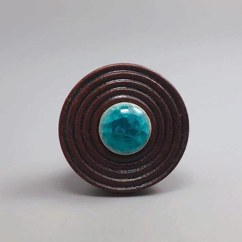 May include: A round, dark brown wooden object with concentric circles. The center features a faceted, teal-colored gemstone. The object is likely a decorative element or a piece of jewelry.