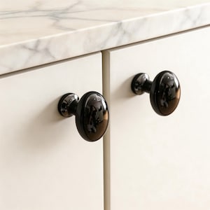 May include: Close-up of two black oval cabinet knobs with a glossy finish. The knobs are mounted on a white cabinet door, with a marble countertop in the background. The knobs are approximately 2 inches in length.