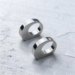 May include: Two polished, silver napkin rings. Each ring has a flat, rectangular base with a rounded, oval opening. The rings are reflective, with a smooth, metallic finish. The background is a light grey, textured surface.