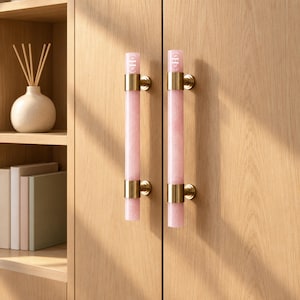 Peut inclure: Deux poignées de placard cylindriques composées de barres en pierre rose clair avec des fixations en métal doré. Chaque poignée est fixée par deux supports dorés et présente un petit emblème circulaire avec les lettres SHN en haut.