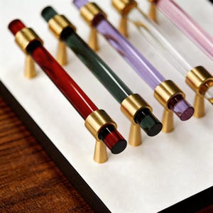 Könnte beinhalten: Eine Auswahl an farbigen Schrankgriffen. Jeder Griff hat einen transparenten, zylindrischen Griff mit farbigem Ende, darunter Rot, Grün, Lila und Rosa. Die Griffe sind mit goldfarbenen Metallringen akzentuiert und auf goldfarbenen konischen Sockeln montiert.