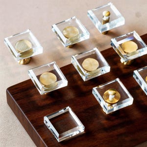 May include: Clear square cabinet knobs with brass accents are arranged on a dark wood surface. The knobs have a modern design, with a clear, square body and a circular brass accent in the center. The knobs are mounted on brass stems.