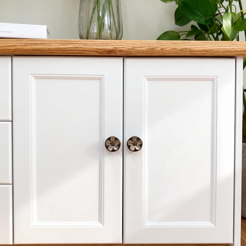 May include: White cabinet doors with flower-shaped knobs. The cabinet has a light wood top. The knobs are dark with a floral design. A vase with flowers sits on top. The cabinet is part of a larger piece of furniture.