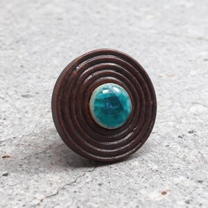 May include: A round, brown wooden ring with concentric circles. The center features a turquoise-colored, faceted gemstone. The ring is set against a neutral gray background, highlighting the contrast between the wood and the stone.