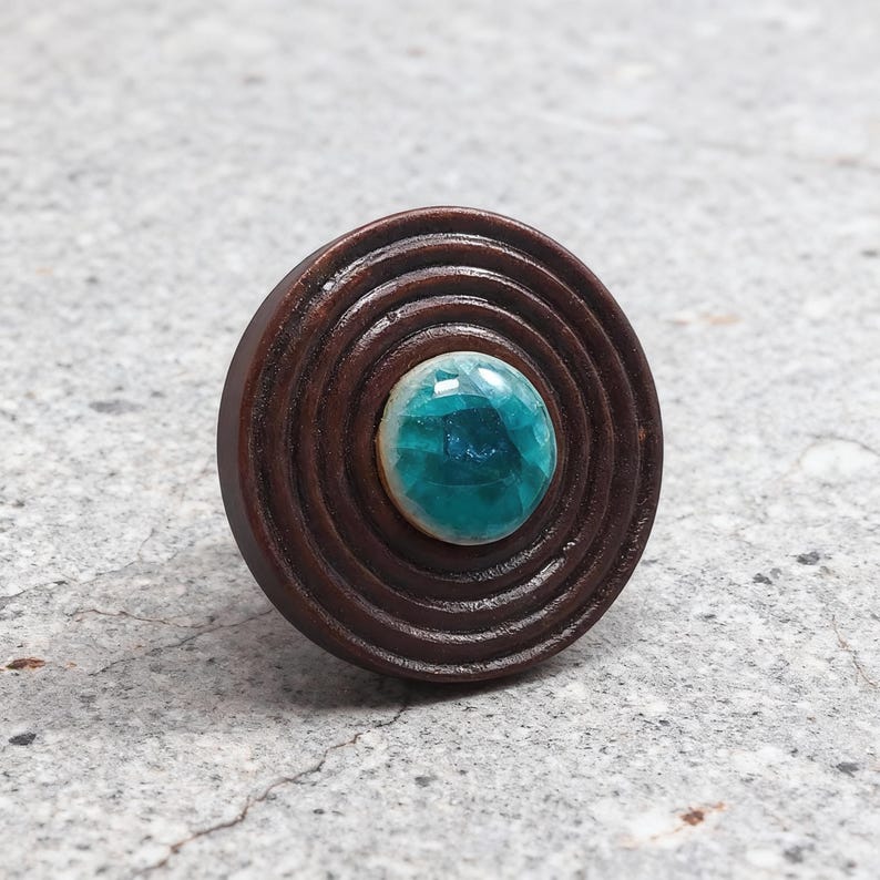 May include: A round, brown wooden ring with concentric circles. The center features a turquoise-colored, faceted gemstone. The ring is set against a neutral gray background, highlighting the contrast between the wood and the stone.