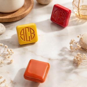 May include: Three square-shaped decorative knobs with a glossy finish. One knob is bright yellow, one is deep red, and one is burnt orange. The yellow and red knobs feature a circular embossed monogram design with the letters ACB in the center.
