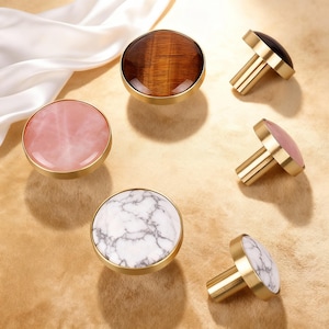 May include: A selection of cabinet knobs with different stone and marble designs. The knobs have a brass finish and include rose quartz, tiger's eye, and white marble with grey veining. The knobs are round and have a simple, elegant design, suitable for various furniture.
