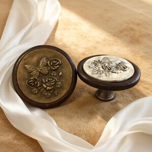 May include: Two decorative cabinet knobs. One is round with a bronze butterfly and rose design. The other is oval with a silver butterfly and rose design. Both have dark brown wooden bases and are set against a neutral background.