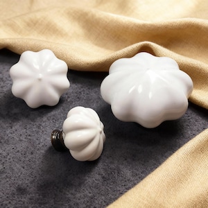May include: Three white ceramic cabinet knobs with a flower-like design. The knobs are glossy and have a smooth surface. One knob is attached to a dark metal base. The knobs are arranged on a gray surface with a tan fabric in the background.