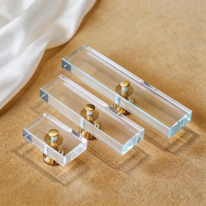 May include: Three clear acrylic cabinet pulls with brass hardware. The pulls are rectangular and vary in size, with a small, medium, and large option. The brass hardware adds a touch of elegance to the modern design. These pulls are ideal for drawers and cabinets.