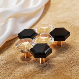 May include: A cluster of six decorative cabinet knobs. The knobs feature a hexagonal design with a clear, faceted center and a black top. The base is a gold-colored metal. The knobs are arranged on a textured, tan surface.