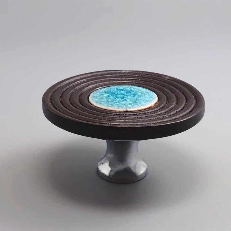 May include: A decorative cabinet knob with a dark brown, circular design and a light blue center. The knob is mounted on a silver-colored base, suitable for furniture or home decor projects.