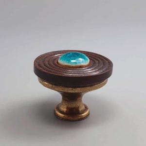 May include: A decorative cabinet knob with a brown, ridged top and a turquoise center. The knob has a brass base and a unique, handcrafted appearance. The knob is approximately 2 inches in diameter and 2 inches tall.