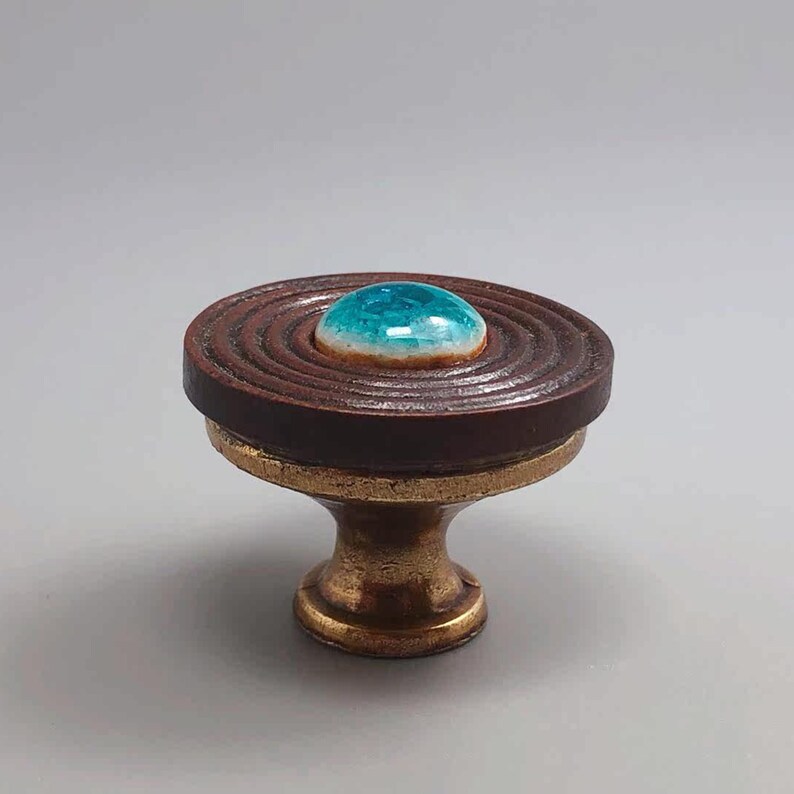 May include: A decorative cabinet knob with a brown, ridged top and a turquoise center. The knob has a brass base and a unique, handcrafted appearance. The knob is approximately 2 inches in diameter and 2 inches tall.