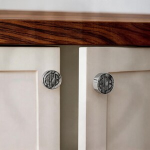 May include: Two circular cabinet knobs with a metallic finish, each featuring a dark, marbled inlay with the engraved monogram letters A, C, and B centered in a classic serif font. The knobs are mounted on light-colored, flat-panel cabinet doors.