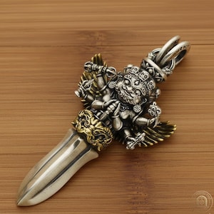May include: A silver and gold-toned metal pendant shaped like a phurba dagger. The handle features a detailed deity figure with multiple arms holding small ritual objects, flanked by wings. A large, smooth, pointed blade extends from the base.