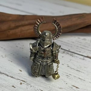 May include: A small metallic knight figurine featuring a silver-toned helmet with large, curved, copper-coloured crest wings. The armour displays a central cross emblem and the figure holds a small, dangling golden ball on a short chain.