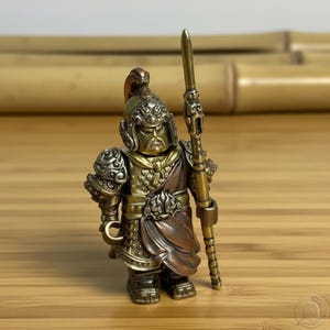 Può includere: Una statuetta metallica di un guerriero che indossa un elmo ornato e un'armatura dettagliata, caratterizzata da un'espressione facciale severa. La figura tiene una lancia lunga e sottile in una mano ed è realizzata con finiture color ottone e rame.