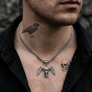 May include: Silver-toned pendant necklace featuring a skull with bat wings and an eye. The necklace is on a chain. A raven tattoo is on the neck, and a skull tattoo is on the chest. The person is wearing a black shirt.
