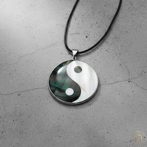 May include: A circular pendant featuring a yin and yang symbol crafted from iridescent dark green and white shell material. The pendant is encased in a silver-toned metal frame and hangs from a thin black cord necklace, creating a balanced design.